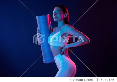 Young woman in sportive clothes is in the studio with neon lights. Holding mat 125079326