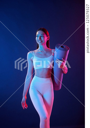 Young woman in sportive clothes is in the studio with neon lights. Holding mat 125079327