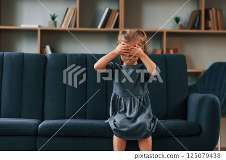 Cute little girl is sitting on the sofa and covering her eyes by the hands Cute little girl is sitting on the sofa and covering her eyes by the hands 125079438