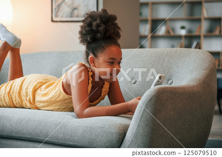 Laying down on sofa with smartphone in hands. Cute black girl in casual clothes is at home at daytime Laying down on sofa with smartphone in hands. Cute black girl in casual clothes is at home at daytime 125079510