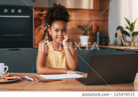 With laptop and paper for drawing. Cute black girl in casual clothes is at home at daytime With laptop and paper for drawing. Cute black girl in casual clothes is at home at daytime 125079515