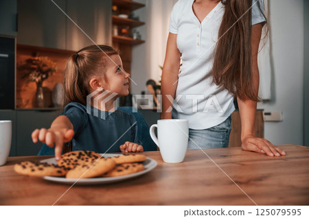 Woman is standing, girl is sitting by the table with cookies. Young mother with her daughter is together at apartment Woman is standing, girl is sitting by the table with cookies. Young mother with her daughter is together at apartment 125079595