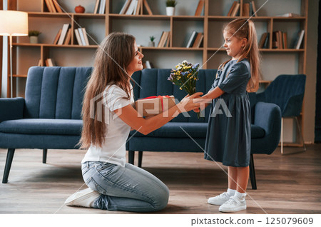 Girl is giving beautiful flowers to a woman. Young mother with her daughter is together at apartment 125079609