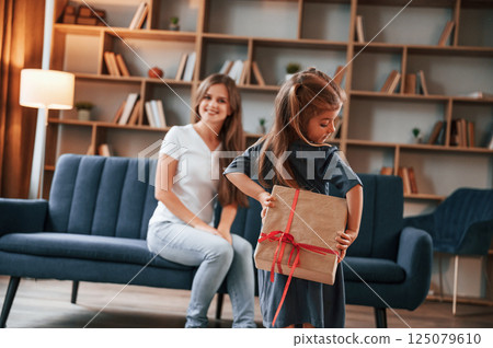 Girl holding gift box behind her. Happy holidays. Young mother with her daughter is together at apartment 125079610