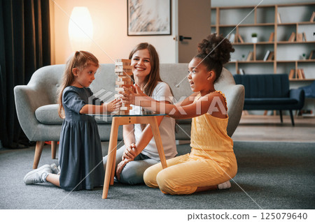 On the floor together. A young woman with two girls is playing a wooden tower game indoors On the floor together. A young woman with two girls is playing a wooden tower game indoors 125079640