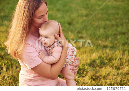 Sitting and kissing the baby. Mother with her newborn son is outdoors on the lawn at the daytime Sitting and kissing the baby. Mother with her newborn son is outdoors on the lawn at the daytime 125079731