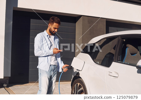 With smartphone. Young stylish man is with electric car at daytime With smartphone. Young stylish man is with electric car at daytime 125079879