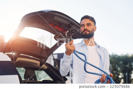 Holding charging cable. Young stylish man is with electric car at daytime 125079886