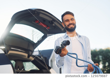 Holding charging cable. Young stylish man is with electric car at daytime 125079887