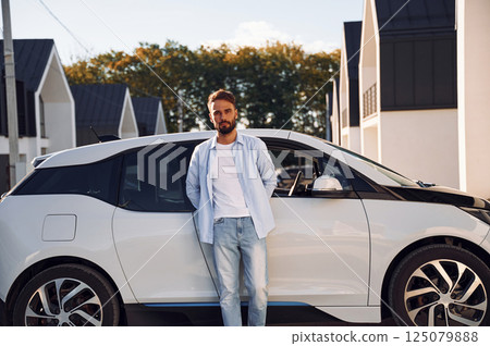 Standing and leaning on the vehicle. Posing for the camera. Young stylish man is with electric car at daytime Standing and leaning on the vehicle. Posing for the camera. Young stylish man is with electric car at daytime 125079888