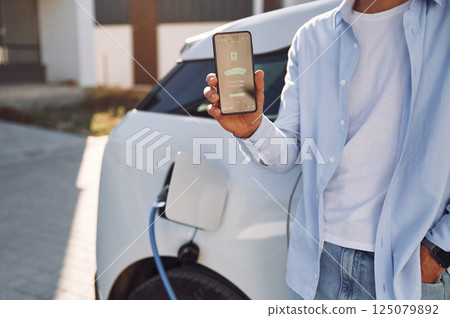 Process of charging is showing on smartphone. Close up view of man with his electric car Process of charging is showing on smartphone. Close up view of man with his electric car 125079892