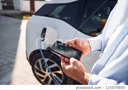Process of charging is showing on smartphone. Close up view of man with his electric car 125079893