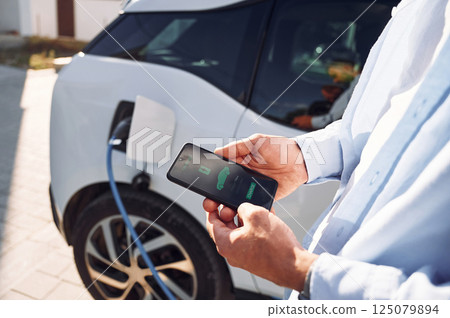 Process of charging is showing on smartphone. Close up view of man with his electric car 125079894