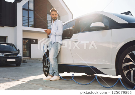 Standing with smartphone and waiting for charge to finish. Young stylish man is with electric car at daytime 125079895