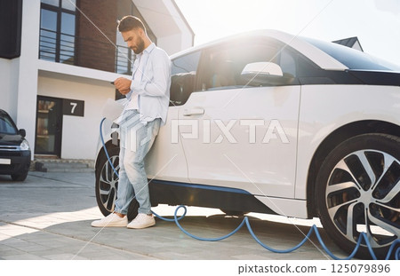 Standing with smartphone and waiting for charge to finish. Young stylish man is with electric car at daytime 125079896