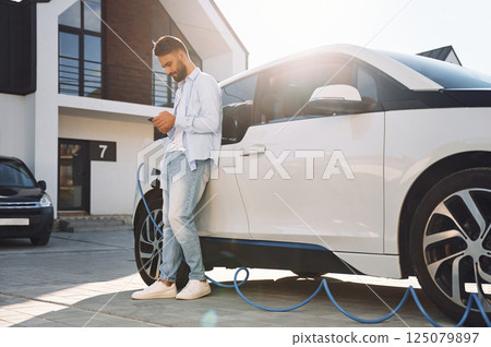 Standing with smartphone and waiting for charge to finish. Young stylish man is with electric car at daytime 125079897