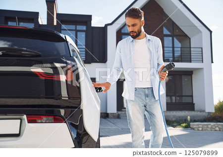 Process of charging the vehicle. Young stylish man is with electric car at daytime Process of charging the vehicle. Young stylish man is with electric car at daytime 125079899