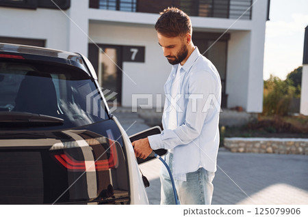 View from the side. Charging the vehicle. Young stylish man is with electric car at daytime View from the side. Charging the vehicle. Young stylish man is with electric car at daytime 125079906