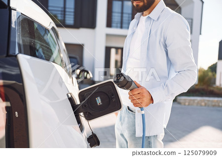 View from the side. Charging the vehicle. Young stylish man is with electric car at daytime View from the side. Charging the vehicle. Young stylish man is with electric car at daytime 125079909