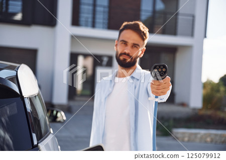 Holding and showing charging cable. Young stylish man is with electric car at daytime 125079912