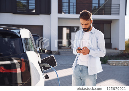 Waiting for charging complete, holding smartphone. Young stylish man is with electric car at daytime Waiting for charging complete, holding smartphone. Young stylish man is with electric car at daytime 125079921