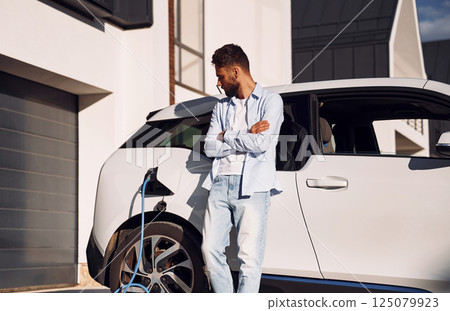 Standing with arms crossed and charging the vehicle. Young stylish man is with electric car at daytime 125079923