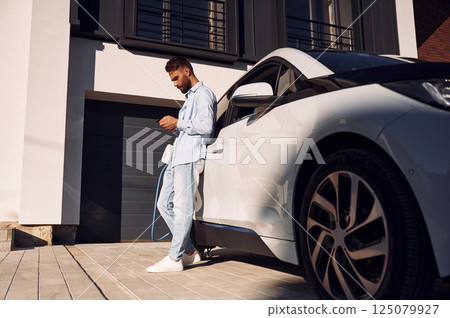 Sunny weather. Holding smartphone and charging the vehicle. Young stylish man is with electric car at daytime 125079927