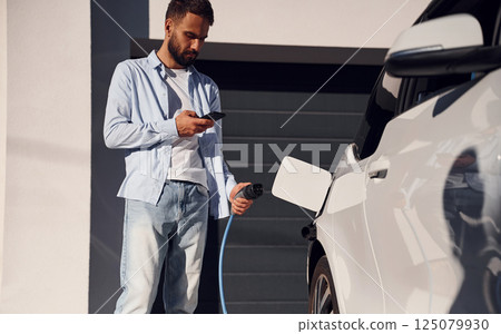 Sunny weather. Holding smartphone and charging the vehicle. Young stylish man is with electric car at daytime 125079930