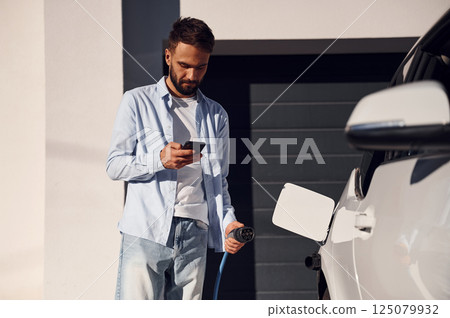Sunny weather. Holding smartphone and charging the vehicle. Young stylish man is with electric car at daytime 125079932