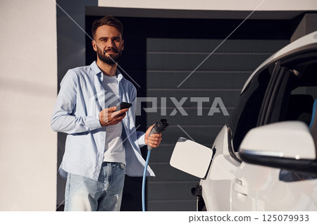 Smartphone and charging cable is in the hands. Young stylish man is with electric car at daytime 125079933