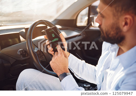Monitoring charging procces by using smartphone. Young stylish man is with electric car at daytime Monitoring charging procces by using smartphone. Young stylish man is with electric car at daytime 125079947