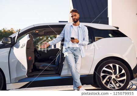 Welcoming to the vehicle. Young stylish man is with electric car at daytime Welcoming to the vehicle. Young stylish man is with electric car at daytime 125079955