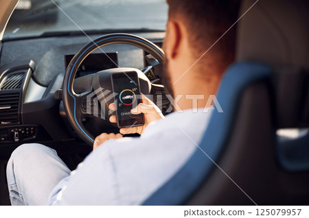 Sitting inside the vehicle, monitoring charging process by using smartphone. Young stylish man is with electric car Sitting inside the vehicle, monitoring charging process by using smartphone. Young stylish man is with electric car 125079957