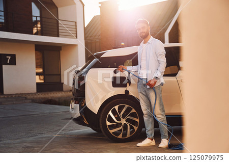 Conception of ecology. Standing with charging cable. Young stylish man is with electric car at daytime 125079975