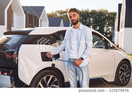 Plugging in charging cable. Young stylish man is with electric car at daytime Plugging in charging cable. Young stylish man is with electric car at daytime 125079976