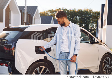 Conception of ecology. Standing with charging cable. Young stylish man is with electric car at daytime Conception of ecology. Standing with charging cable. Young stylish man is with electric car at daytime 125079977