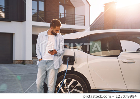 Charging the vehicle near the house. Holding smartphone. Young stylish man is with electric car at daytime Charging the vehicle near the house. Holding smartphone. Young stylish man is with electric car at daytime 125079984
