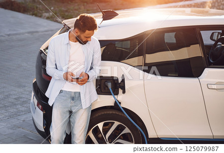 Waiting for the charge completes. With smartphone in hands. Young stylish man is with electric car at daytime 125079987