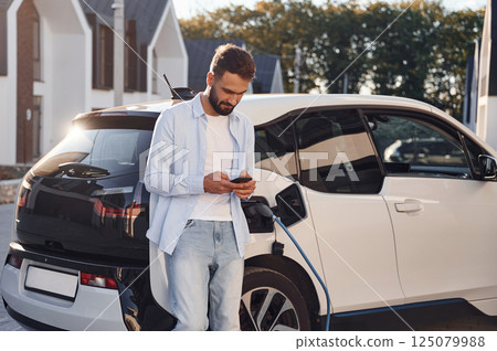 Waiting for the charge completes. With smartphone in hands. Young stylish man is with electric car at daytime 125079988
