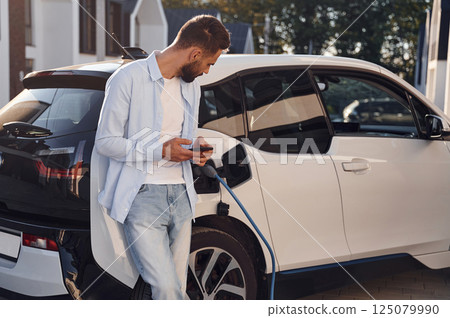 Beautiful young stylish man is with electric car at daytime charging the vehicle and holding smartphone 125079990