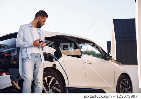 Standing and looking at vehicle that is being charging. Young stylish man is with electric car at daytime Standing and looking at vehicle that is being charging. Young stylish man is with electric car at daytime 125079997