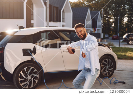 Smiling and having fun while vehicle is being charged. Young stylish man is with electric car at daytime Smiling and having fun while vehicle is being charged. Young stylish man is with electric car at daytime 125080004