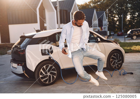 Jumping and having fun. Charging the vehicle. Young stylish man is with electric car at daytime Jumping and having fun. Charging the vehicle. Young stylish man is with electric car at daytime 125080005