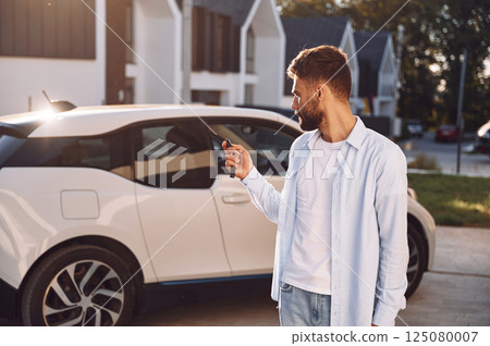Standing and holding the keys. Young stylish man is with electric car at daytime Standing and holding the keys. Young stylish man is with electric car at daytime 125080007