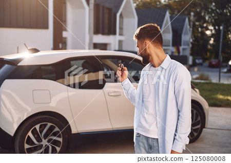 Standing and holding the keys. Young stylish man is with electric car at daytime 125080008