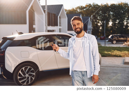 Standing and holding the keys. Young stylish man is with electric car at daytime Standing and holding the keys. Young stylish man is with electric car at daytime 125080009