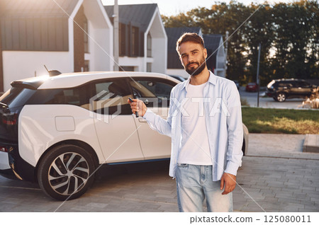 Standing and holding the keys. Young stylish man is with electric car at daytime 125080011
