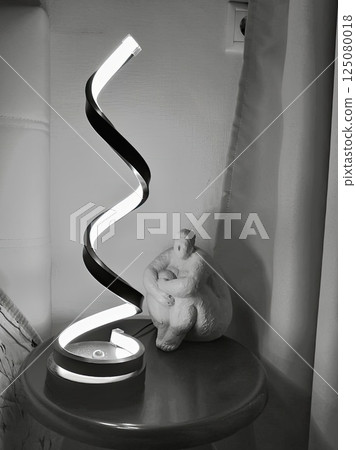 Bw photo of a spiral lamp and a ceramic figure sitting in contemplation. Minimalistic night interior with soft lighting and a calm meditative mood. Great for backgrounds, mindfulness concepts Bw photo of a spiral lamp and a ceramic figure sitting in contemplation. Minimalistic night interior with soft lighting and a calm meditative mood. Great for backgrounds, mindfulness concepts 125080018