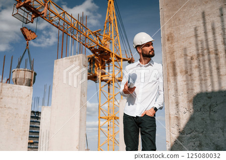Standing and holding plan in hands. Man in uniform is working on the construction site 125080032