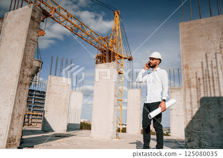 Talking by phone and holding document in hands. Man in uniform is working on the construction site 125080035